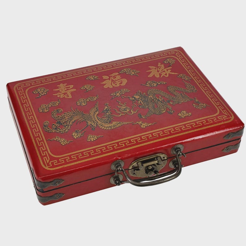 May include: A red rectangular box with gold Chinese characters and illustrations of a dragon and phoenix. The box has a metal clasp and handle, and a decorative border.