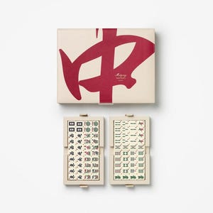 Puede incluir: Un juego de Mahjong beige con un gr&aacute;fico rojo en la tapa y dos porta-fichas abiertos. Los porta-fichas muestran fichas con varios s&iacute;mbolos y caracteres en verde, rojo y negro. La caja tiene el texto "Mahjong" en rojo.