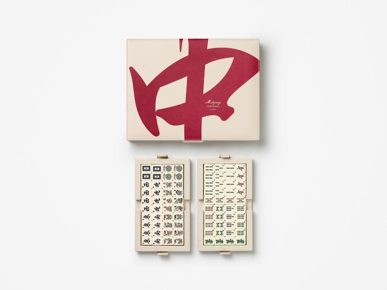 Puede incluir: Un juego de Mahjong beige con un gr&aacute;fico rojo en la tapa y dos porta-fichas abiertos. Los porta-fichas muestran fichas con varios s&iacute;mbolos y caracteres en verde, rojo y negro. La caja tiene el texto "Mahjong" en rojo.