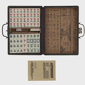 May include: An open Mahjong set in a black case, featuring tiles with Chinese characters and symbols. The set includes dice and a booklet with instructions. The case is open, revealing the organized arrangement of tiles.