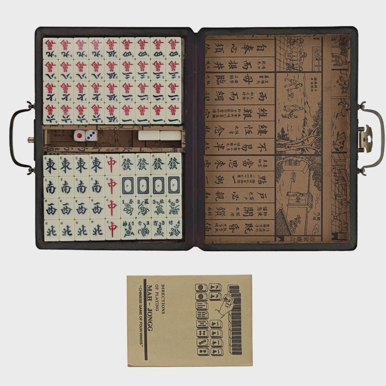 May include: An open Mahjong set in a black case, featuring tiles with Chinese characters and symbols. The set includes dice and a booklet with instructions. The case is open, revealing the organized arrangement of tiles.