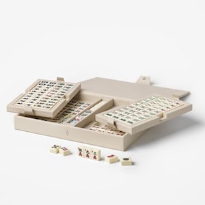 Puede incluir: Un juego de Mahjong beige con fichas y una caja de almacenamiento. La caja est&aacute; abierta, revelando m&uacute;ltiples bandejas de fichas con s&iacute;mbolos negros y rojos. Varias fichas sueltas est&aacute;n esparcidas frente a la caja.