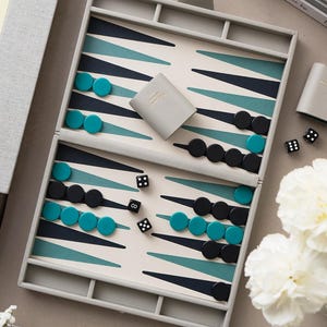 May include: A backgammon set with a grey case, teal and black game pieces, and dice. The game board features a cream-coloured background with teal and navy blue triangles. A small grey case is also included.