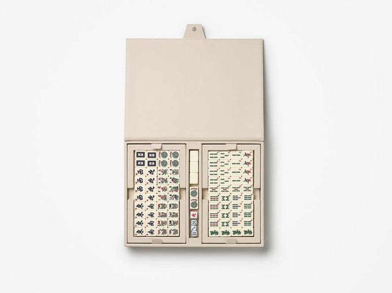 Puede incluir: Un juego de Mahjong beige abierto, que revela fichas y dados. Las fichas est&aacute;n en dos compartimentos dentro de un estuche beige a juego. Las fichas muestran s&iacute;mbolos y caracteres en verde, rojo y negro. El estuche es rectangular.