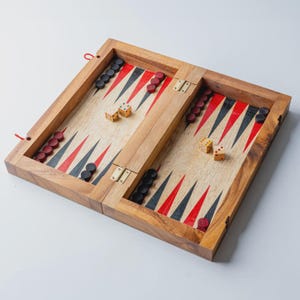 May include: A wooden backgammon set with red, black, and white triangles on the playing surface. The set includes red and black game pieces, dice, and brass hinges. The wooden frame is light brown, and the game is open and ready to play.