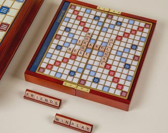 以下が含まれることがあります： 赤と青のグリッドを持つ木製の Scrabble ボードゲームで、「TRAVEL」と「BEST」という文字が書かれています。ゲームは開いており、「FRIENDS」と「WIN PLAY」と書かれた追加のタイルがあります。ボードは木製のケースに入っています。