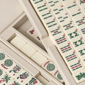 Puede incluir: Primer plano de un juego de Mahjong con fichas de color marfil, dispuestas en un estuche color crema. Las fichas presentan caracteres chinos, n&uacute;meros y s&iacute;mbolos en rojo y verde. Algunas fichas est&aacute;n etiquetadas como "JOKER".