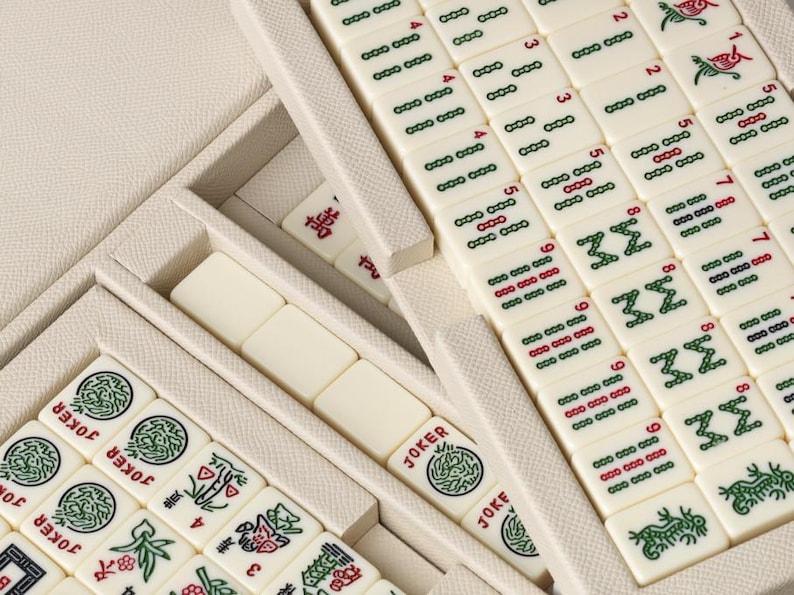 Puede incluir: Primer plano de un juego de Mahjong con fichas de color marfil, dispuestas en un estuche color crema. Las fichas presentan caracteres chinos, n&uacute;meros y s&iacute;mbolos en rojo y verde. Algunas fichas est&aacute;n etiquetadas como "JOKER".