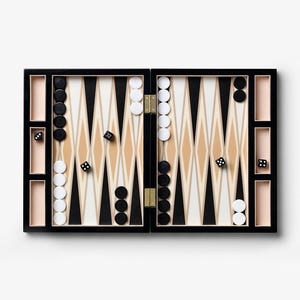 May include: An open backgammon board in black and tan, with a geometric design of triangles in black, white, and tan. The board is set up with black and white game pieces and dice. The board is rectangular and has storage compartments for the pieces and dice.