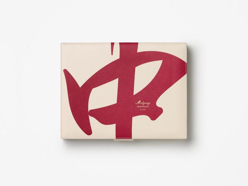 Puede incluir: Un estuche rectangular blanquecino con un gran dise&ntilde;o rojo abstracto. El dise&ntilde;o es una forma caligr&aacute;fica estilizada. El estuche tiene el texto "Mahjong Printworks" en una fuente peque&ntilde;a y elegante. El estuche est&aacute; cerrado.