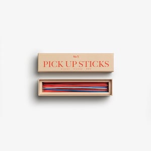 Puede incluir: Una caja beige con la inscripción "No 5 PICK UP STICKS" en letras rojas. Debajo, una caja revela palitos rojos, azules y blancos. Un juego clásico.