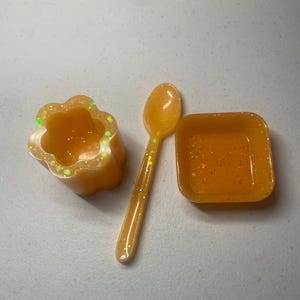 May include: A set of three small orange resin dishes with glitter. The dishes are shaped like a flower, a square, and a spoon.