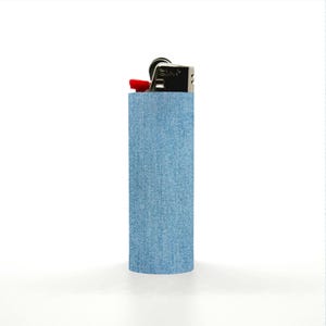 May include: A silver lighter with a red ignition button is encased in a light blue denim sleeve. The denim sleeve is a solid color and covers the majority of the lighter's body. The background is white.