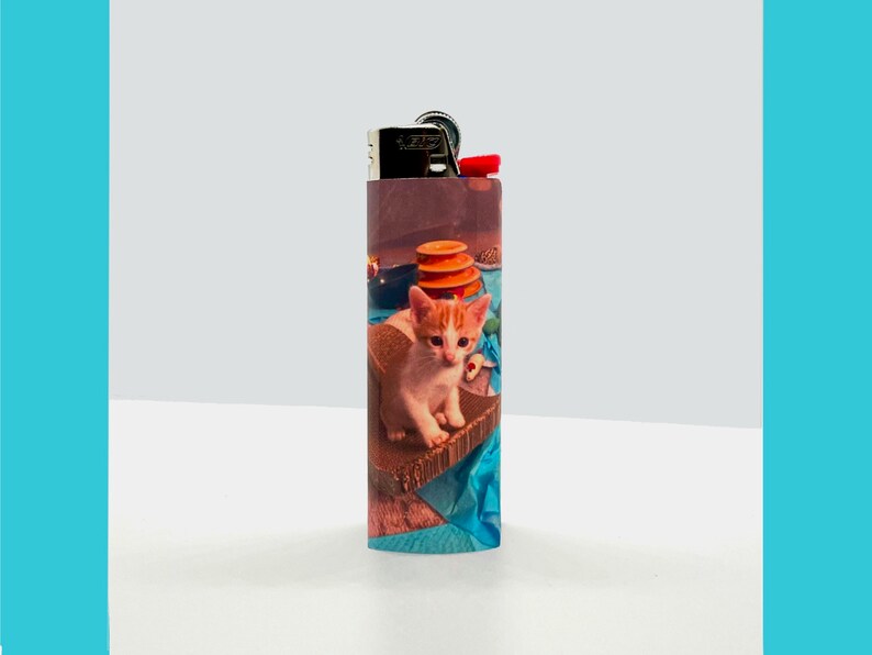 Puede incluir: Un encendedor con un dise&ntilde;o colorido que presenta un gatito sentado en una superficie azul y marr&oacute;n. El gatito tiene pelaje blanco con marcas naranjas.