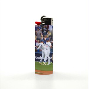 May include: A lighter with a baseball themed design. The image shows two baseball players celebrating a home run. The lighter is silver with a red top and a clear plastic body. The image on the lighter is of a baseball game with players in white and blue uniforms.