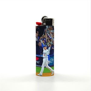 May include: A rectangular lighter with a baseball theme. The lighter features a player in a blue and white uniform, celebrating with a raised arm. The background shows a crowd of cheering fans. The lighter has a black top and a red ignition button.