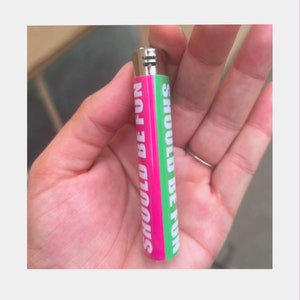 May include: A pink and green lighter with the words "SHOULD BE FUN" printed vertically. The lighter is held in a hand and has a silver metal top.