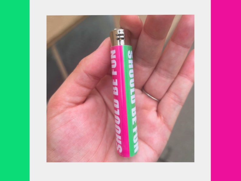 May include: A pink and green lighter with the words "SHOULD BE FUN" printed vertically. The lighter is held in a hand and has a silver metal top.