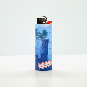 May include: A blue and white lighter with a graphic of a building with the word "SURF" on it. The lighter has a red warning label that says "PROPERTY OF THE SURF BACK ALLEY".
