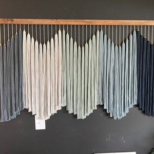 May include: A wall hanging made of yarn with a wooden dowel at the top. The yarn is in shades of blue, gray, and white, and is arranged in a staggered pattern. The yarn is tied to the dowel with gold beads.