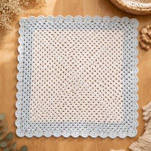 May include: A square crochet blanket featuring a white center with a granny square pattern, surrounded by a light blue border and finished with a scalloped edge. The textile is crafted with a consistent stitch pattern throughout the design.