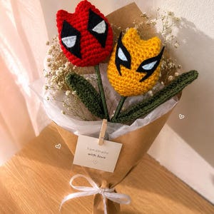 May include: A bouquet of two crocheted flowers with green stems and leaves, wrapped in brown paper with a white ribbon. One flower is red with black and white eye patches, and the other is yellow with black and white eye patches. A small tag reads handmade with love.