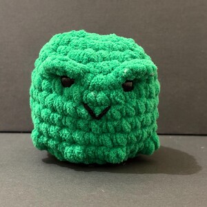 May include: A round, green, crocheted plush toy featuring a textured, yarn-based surface. The object has two small, dark, inset eyes and a simple, black, V-shaped embroidered mouth, giving it a distinct, grumpy facial expression.