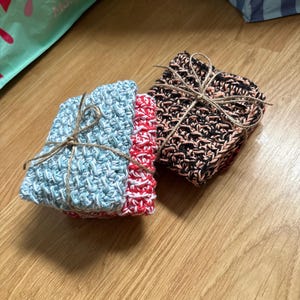 Peut inclure: Deux piles de carrés de débarbouillettes crochetés, attachés avec de la ficelle. Une pile présente des bords bleu clair et rouges, tandis que l'autre a des tons noirs et pêche. Les débarbouillettes sont faites de fil texturé.