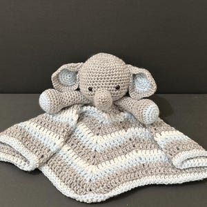 May include: A crochet security blanket featuring a stuffed elephant head and arms attached to a square, chevron-patterned blanket. The yarn is colored in alternating stripes of light gray, white, and pale blue, with black plastic safety eyes.