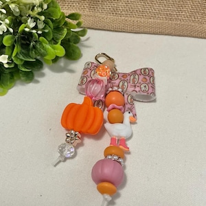 Könnte beinhalten: Ein Schlüsselanhänger mit einer gemusterten rosa Schleife und zwei Perlensträngen. Ein Strang hat eine orangefarbene Kürbisperle und klare facettierte Perlen. Der andere Strang enthält orange und rosa Rundperlen, eine weiße Gans und einen Goldverschluss.
