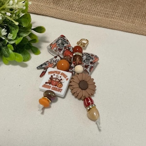 Könnte beinhalten: Ein dekorativer Schlüsselanhänger mit einer Stoffschleife mit Kürbis- und Kreuzmuster, einer quadratischen weißen Perle mit dem Text Pumpkin Spice & Jesus Christ, einer braunen Sonnenblumenperle sowie orangefarbenen, braunen und roten Perlen.