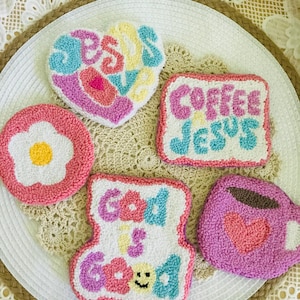 Op de afbeelding: Een verzameling kleurrijke, getextureerde onderzetters met geborduurde ontwerpen. De onderzetters tonen zinnen als "Jesus Loves", "Coffee Jesus" en "God is Good", samen met een bloem en een koffiekopje. Het kleurenpalet omvat roze, blauw, geel en wit.
