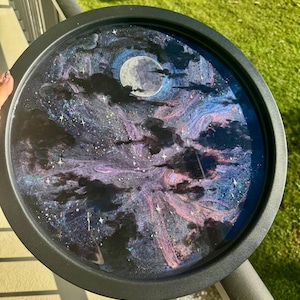 May include: Circular black tray with a cosmic design. It depicts a full moon, dark clouds, and a swirling galaxy in purple, blue, and white. Glitter adds sparkle to the night sky scene.