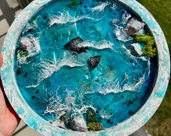 May include: A round, decorative tray featuring a vibrant ocean scene. The tray has a textured, white and blue rim, surrounding a turquoise and blue ocean design with white waves, rocks, and two black stingrays. Green moss adds detail.