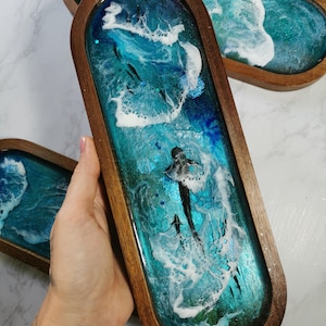 May include: An elongated oval wooden tray featuring a resin inlay that mimics a deep blue ocean scene with white crashing waves and a small black shark figure swimming through the center. The tray has a smooth, dark brown wooden frame.