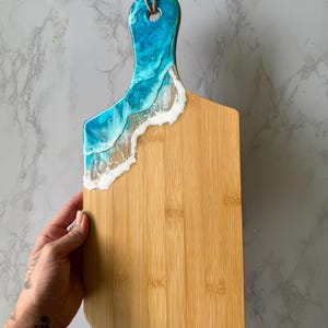 Op de afbeelding: Een bamboe snijplank met een handvat, versierd met een blauw-wit hars golfontwerp. Het handvat heeft een gat om op te hangen, met een natuurlijk touw. De plank is gemaakt van bamboe.