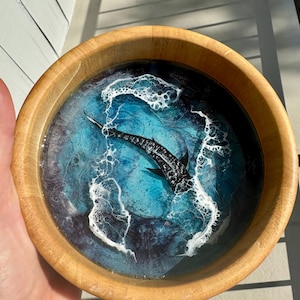 Puede incluir: Cuenco de madera con una escena oceánica de resina. El cuenco presenta un tiburón blanco y negro nadando en olas de resina azul y blanca. El cuenco es de madera clara.