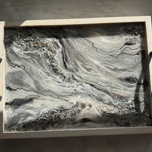 May include: A rectangular serving tray with a light wood frame and black handles. The tray's surface features a swirling design in shades of gray, white, and silver, with black linear accents and embedded glitter and stones.