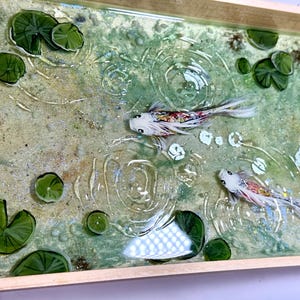 以下が含まれることがあります： 穏やかな池のシーンが描かれた長方形のトレイ。白とカラフルな2匹の鯉が、緑のスイレンの葉の間を泳いでいます。水面はキラキラと輝き、さざ波があり、明るい木製のフレームで囲まれています。