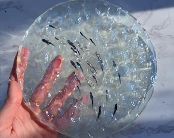 May include: A circular, translucent resin coaster featuring a textured, rippled surface that mimics the appearance of water. Embedded within the clear material are numerous small, dark, slender fish-shaped figures scattered throughout the piece.