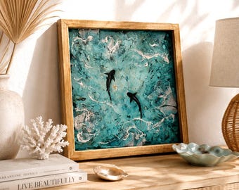 May include: A square wall art piece in a light wood frame featuring an abstract painting of two black sharks swimming in textured turquoise and teal water with white foam accents. The artwork displays a dynamic ocean scene with layered paint.