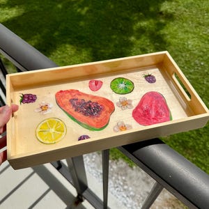 May include: A rectangular wooden serving tray with a light wood finish. The tray is decorated with colorful illustrations of fruit slices, including papaya, watermelon, kiwi, and lemon. Small white flowers and berries are also featured.