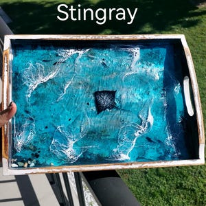 May include: Serving tray with a marine theme. The tray has a turquoise and blue ocean design with white wave details and a black stingray. The tray has a white frame and handles, with the word "Stingray" in white.
