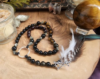 May include: A beaded necklace featuring black, garnet, and tiger eye stones interspersed with small metallic pyrite nuggets. The necklace includes a silver-toned circular ring, a silver-toned pentagram charm, and a brown and white patterned feather.