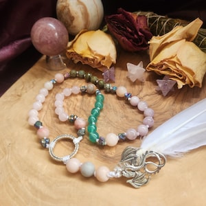 May include: A beaded necklace with a silver dragon charm and a white feather. The necklace is made with pink, green, and brown beads. The necklace is on a wooden surface with dried flowers and other decorative items.