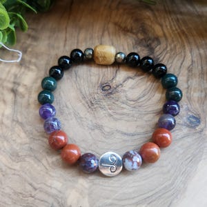 May include: A beaded bracelet featuring a variety of colorful gemstones, including black, green, purple, and red-brown beads. A silver charm with a symbol is centered on the bracelet. A wooden bead and other metallic beads are also included.