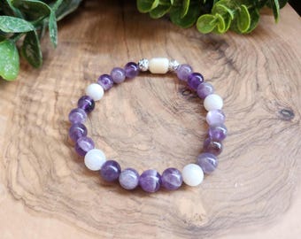 May include: A beaded bracelet featuring alternating purple and white gemstone beads. The bracelet has a central rectangular bead and silver accents. The bracelet is displayed on a wooden surface with green foliage in the background.