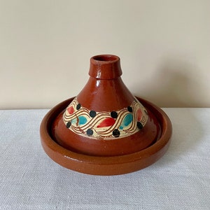 May include: A brown ceramic tagine pot, featuring a conical lid and a broad, shallow base. The lid is adorned with a decorative band of red, blue, and black patterns. This tagine is designed for cooking and serving traditional Moroccan cuisine.