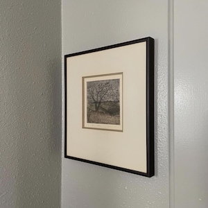 May include: Framed artwork with a detailed tree illustration. The artwork is in a black frame with a white mount, mounted on a light grey wall. The artwork's style appears to be a pen and ink drawing.