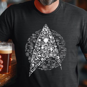 May include: Black t-shirt with a white Starfleet emblem design. The emblem is made up of numerous small, detailed illustrations. The person in the image is holding a pint of beer.